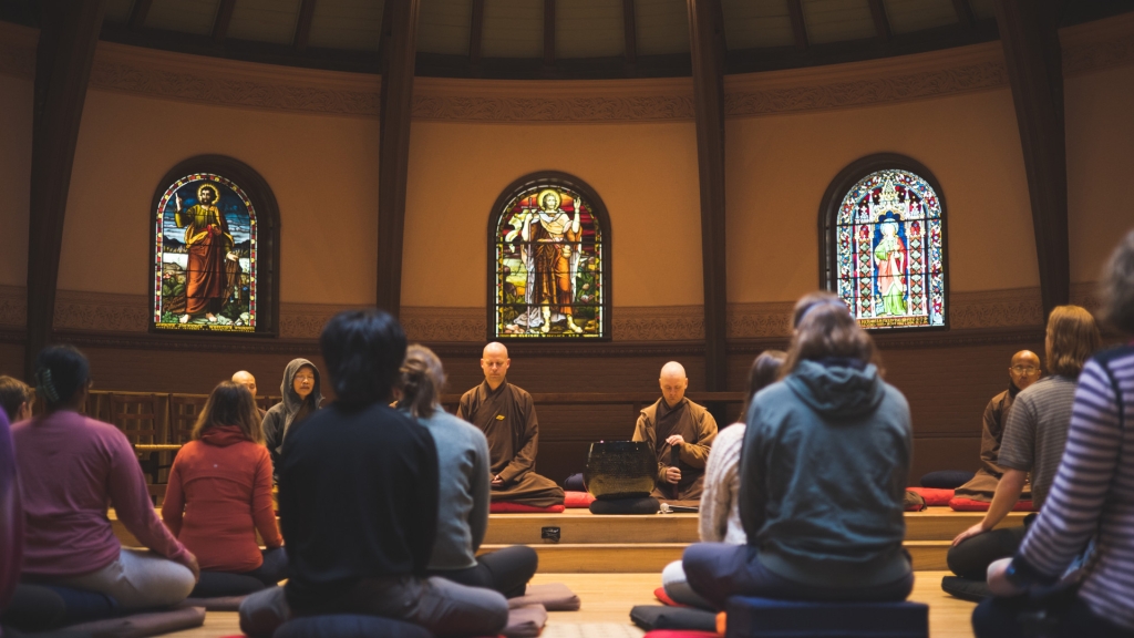 Two rows of students and community members sit on floor cushions, participating in a silent meditation led by Buddhist monastics visiting Dartmouth from monasteries in California and Mississippi. The meditation is taking place in the calm environment of Rollins Chapel, where three stained glass windows in the background depict scenes from the Christian religious tradition.