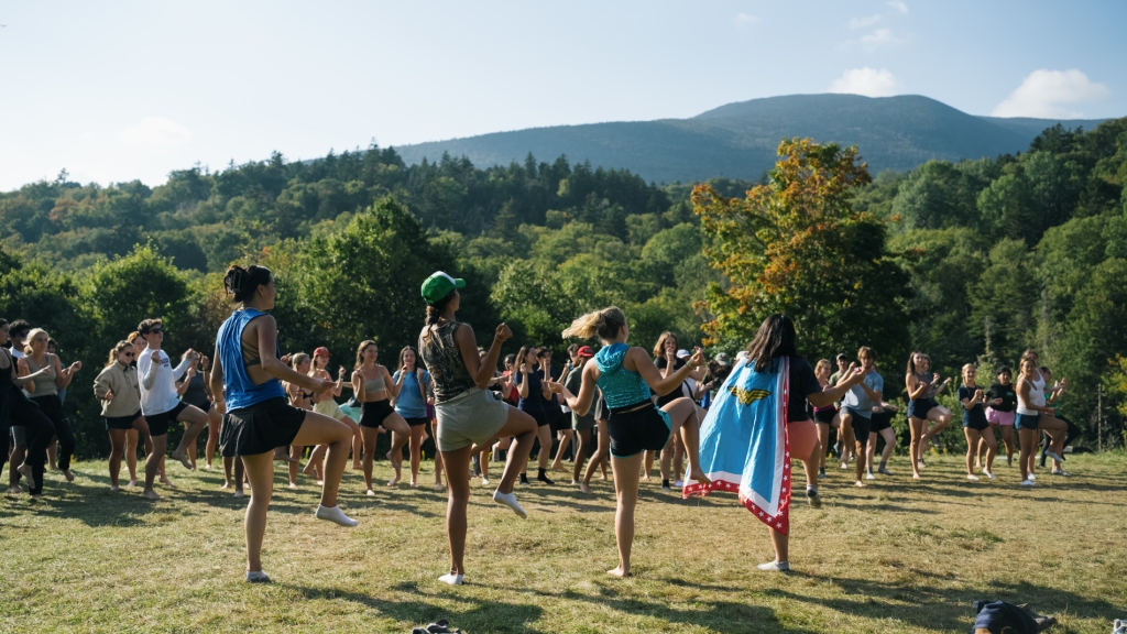 On a sunny day in September, about 35 undergraduate students participate in a First Year Trips activity on a grassy knoll, with lush green trees and a tree-covered mountain in the background. Four student leaders, including one who is wearing a Wonder Woman cape and another who is wearing a sparkly shirt, lead them in a lighthearted group dance that involves playfully swaying their arms and stepping with their legs.